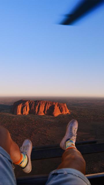 Point of view of person looking down at Uluru from a helicopter. Person's legs are hanging over